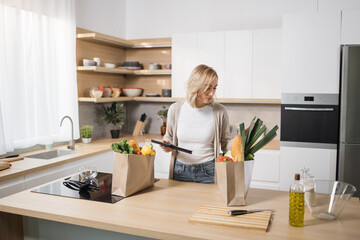 Confident serious woman using tablet preparing vegetable salad in kitchen. Young caucasian female chef searching recipes, ordering food online, watching cooking videos preparing healthy food at home.