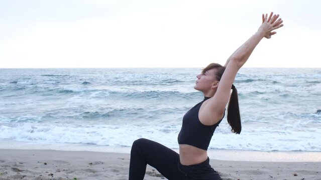 A caucasian young woman in a black sports suit doing hands-up yoga asana on the ocean beach
