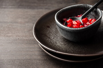 Overhead view of black bowl with pomegranate seeds and spoon on plates and dark table, horizontal, with copy space