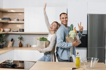 Positive joyful young indian man and blond woman standing together in kitchen, smiling, standing back to back and unpacking grocery paper bag. Concept of family, domestic lifestyles and healthy eating