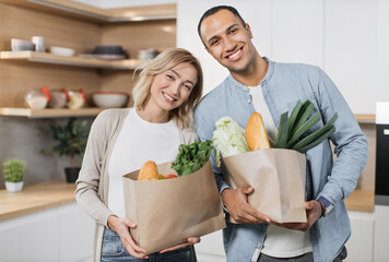 Cheerful young indian man and woman standing together on kitchen, smiling, looking at camera and unpacking grocery paper bag. Concept of family, domestic lifestyles and healthy eating.