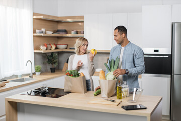 Positive young people in love unpacking shopping back with fresh organic products. Caucasian family standing together on kitchen and enjoying domestic chores.