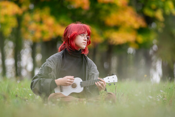 Naklejka na ściany i meble A girl with red hair plays a white ukulele. Autumn park with yellow foliage.