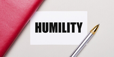 On a light gray background, there is a golden pen, a burgundy notebook and a white blank card with a place to insert text HUMILITY. Business concept.