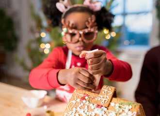 Girl decorating gingerbread house. Kids assembling  tasting gingerbread cookies for Christmas holidays. Happy moments at home concept