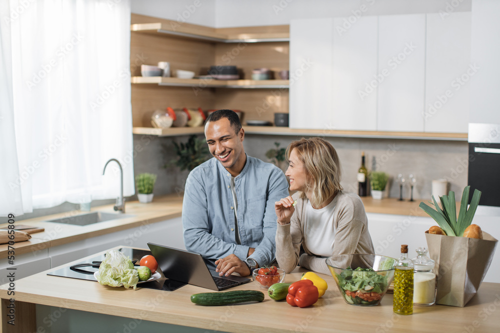 Wall mural Happy young multiracial couple using laptop computer preparing healthy food diet vegetable salad at home together, searching recipes, ordering shopping online, watching cooking class in kitchen.