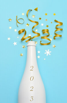 New Years 2023 Concept. White Champagne bottle on a teal blue background with gold stars and ribbon and the date 2023 on the bottle.