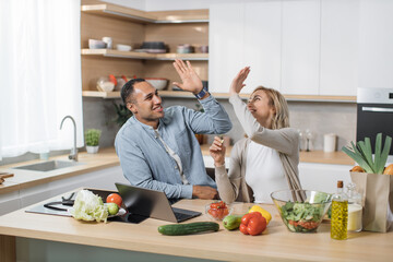 Excited joyful young multiracial couple using laptop computer preparing healthy food diet vegetable...