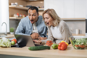 Handsome excited indian man and caucasian blond joyful woman having good news using laptop searching recipes, ordering shopping online, watching cooking class during preparing delicious vegan salad