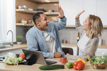Excited joyful young multiracial couple using laptop computer preparing healthy food diet vegetable...