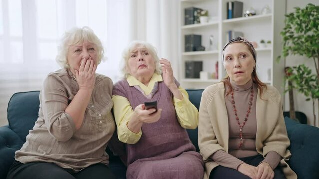 Shocked women in their 60s watching TV sitting on sofa, bad news, horror film