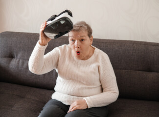 An elderly woman is very surprised by what she sees while playing using virtual reality glasses. Modern technologies. A new experience in the life of an old person. The idea of original gift 