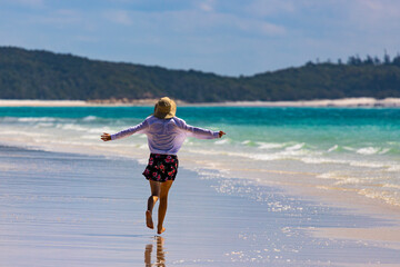 Fototapeta na wymiar a beautiful girl in a dress, shirt and hat spreads her arms in joy as she walks along whitehaven beach; leisure on whistundays islands, queensland, australia; famous white sand dunes
