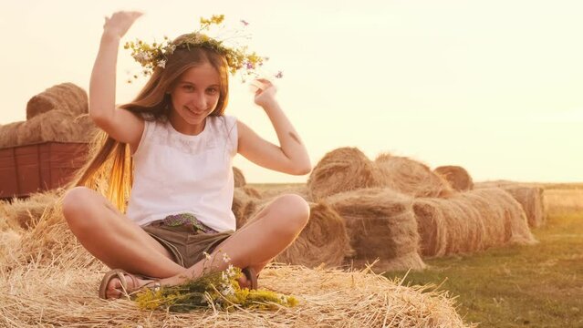 Beautiful ukrainian long hair teen girl sitting on hay stack in the field, weatring flower wreath and smiling. Pretty female teenager in countryside in sunset time