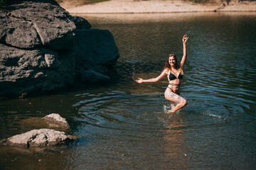 Fototapeta premium A young girl with long blond hair, a slender figure, in a black swimsuit jumps in the river against the backdrop of rocks and laughs.