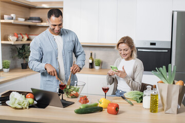 Portrait of loving multinational couple cooking salad together on a kitchen. Cheerful blond woman using mobile phone to find recipe of vegan salad while her husband mixing vegetables in a glass bowl