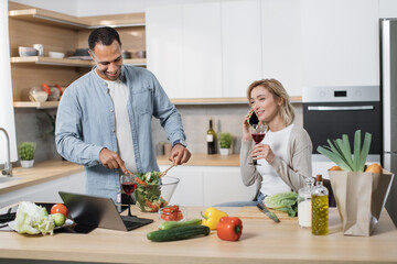 Cheerful blond woman using mobile phone to find recipe of vegan salad while her husband mixing vegetables in a glass bowl, cooking salad together on a kitchen and drinking red wine.