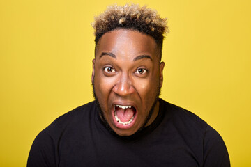 An outraged African-American yells with all his might in outrage close-ups on camera on a yellow background. An enraged black guy screams loudly with his mouth wide open and bulging eyes. 
