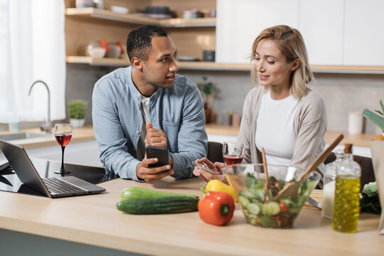 Happy multinational couple using cell phone while having breakfast at kitchen, positive man and woman eating healthy food, checking photos on social media and smiling, having video conference