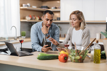 Happy multinational couple using cell phone while having breakfast at kitchen, positive man and...