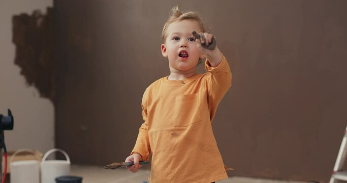 Smiling two-year-old boy stands in the room and holds brush in hands. The blond boy is wearing orange sweater, arm and sweater are smeared with brown paint. Behind is painted wall and buckets of paint