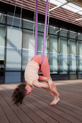 Sporty woman doing aero yoga in hammock. Brunette girl in sportswear exercising aerial yoga on terrace. Windows in background