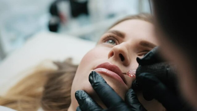 Beautiful girl lying while master making permanent makeup for her lips in salon. Closeup view of face