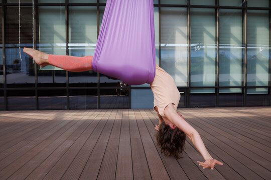 Sporty woman doing aero yoga in hammock. Brunette girl in sportswear exercising aerial yoga on terrace. Windows in background