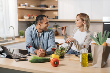 Cheerful married multinational couple using laptop while cooking healthy food in kitchen, blond young wife spouses having fun while feeding her husband wooden spoon with salad from fresh vegetables