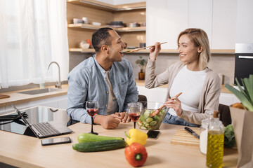 Cheerful married multinational couple using laptop while cooking healthy food in kitchen, blond...