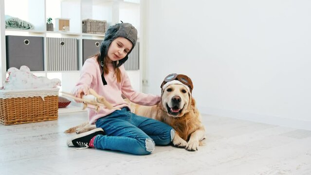 Little girl wearing pilot hat playing with toy plain and golden retriever dog in pilot glasses lying next to her