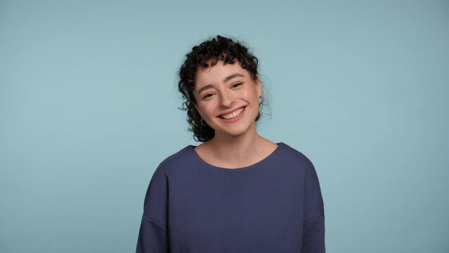 Close up laughing happy young curly woman in blue sweater posing looks camera. Portrait smiling positive beautiful female standing on isolated light blue background. People emotions lifestyle concept