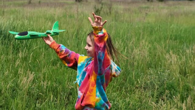 Preteen girl child wearing colorful hoodie playing with toy plane in the field with green grass and smiling. Pretty young female kid launching airplane at the nature