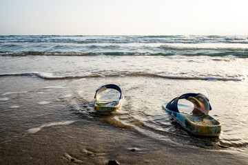 focused colorful and sandy kids slippers sandals is on the beach, the sea and beach is blurred in the background, sunset and wavy sea