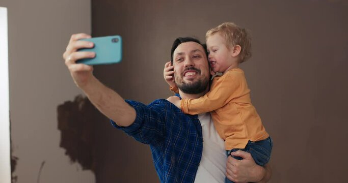 Man with dark hair and beard holds little blond son in arms. Boy in orange sweater hugs dad. Man in blue shirt is holding phone and taking selfie with son. They are having fun and laughing.