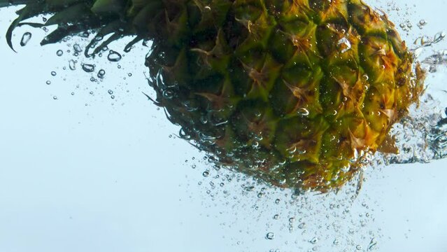 Whole sweet pineapple fall into clear water in super slow motion close up.
