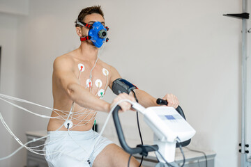 Man athlete with breath mask and electrodes training on bike simulator, examining his...