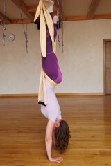 Young woman in sportswear practices different inversion aerial yoga with a hammock in beige hall....