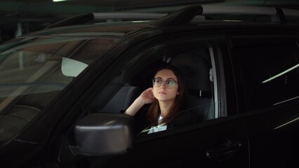 Plakat A business woman looks in a car mirror and straightens her hair. A young woman in glasses and a suit in a parking lot sits in a car and straightens her hair.