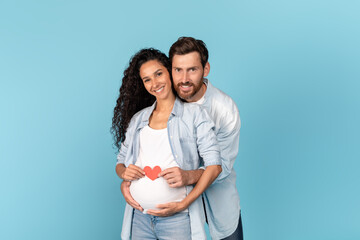 Happy millennial caucasian man with beard hugging pregnant arabic woman, holding heart on belly