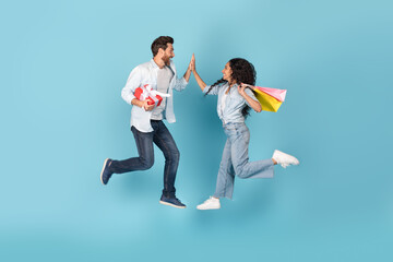 Excited glad millennial international couple with gift and bags jump in air, give high five on copy...