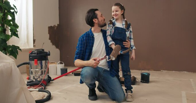 Dad with beard sat down and hugged daughter. He is wearing t-shirt and checkered shirt. Girl is dressed in denim overalls. They are holding rollers, looking at the camera and smiling.