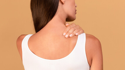 Rear view of lady rubbing her neck, having pain or spasm, beige studio background, cropped