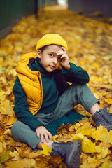 portrait of a fashionable serious child boy autumn sitting on a trail in orange leaves in the afternoon at a green fence on the street.