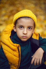 portrait of a fashionable serious child boy autumn sitting on a trail in orange leaves in the afternoon at a green fence on the street.