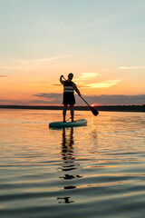 A man in shorts on a SUP board with an oar against the backdrop of a bright sunset sky swims in the...