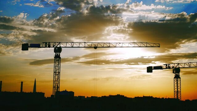 Dark silhouettes working and moving of construction cranes and unfinished residential building on yellow beautiful sunrise and blue sky background. Housing construction, apartment block in city.