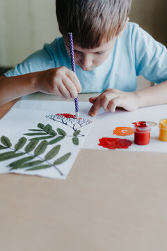 Cute child siting at desk and drawing rowanberries on album sheet with dry rowan leaves
