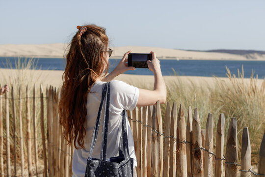 A red haired girl is photographing the sea with her smartphone in front of a sandy beach in south east France. The Dune du Pylat is visible in the background