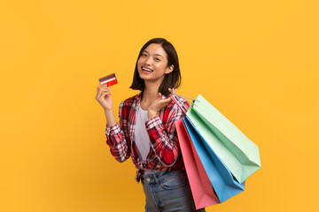 Cashback concept. Happy asian lady posing with credit card and colorful shopping bags over yellow...